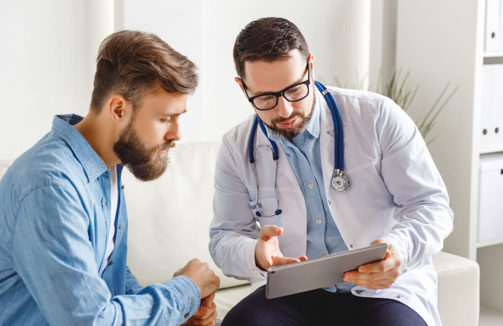 Medical practitioner in glasses showing test results to man and explaining diagnosis while sitting on couch in office of modern clinic